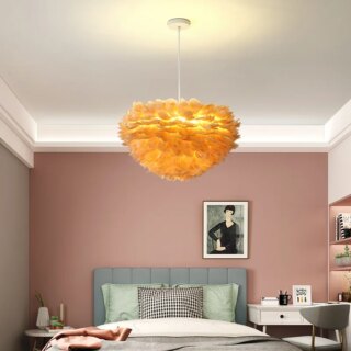 Photo représentant un lustre en plumes en forme de nid orange suspendu dans une chambre d'ado au mur du fond rose poudré, plafond blanc, lit avec trois coussins et petit bureau blanc