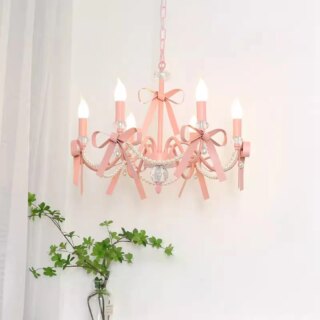 Photo d'un lustre rose chandelier à six ampoules bougies et noeuds roses orné de guirlandes en perles et de cristal en bas des bougeoirs et en bas du corps du lustre, le tout suspendu devant un mur blanc avec un voilage blanc sur la gauche et des branches à feuilles vertes dans un vase transparent